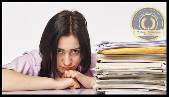 A lady with discouraged expression sitting behind a large pile of paperwork. A Flourishing Life Society article on productivity