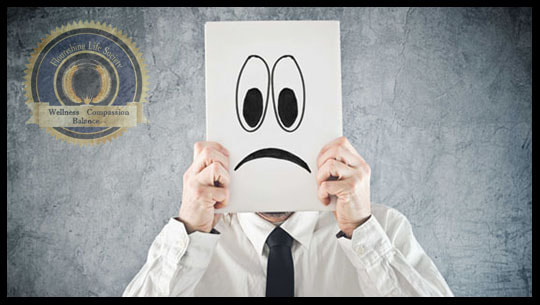 A cartoon frowny face held up by real person. Representing feelings. Emotional regulations is not showing an emotion we're not experiencing. Emotional regulation is harnessing the power of emotions. A Flourishing Life Society Article